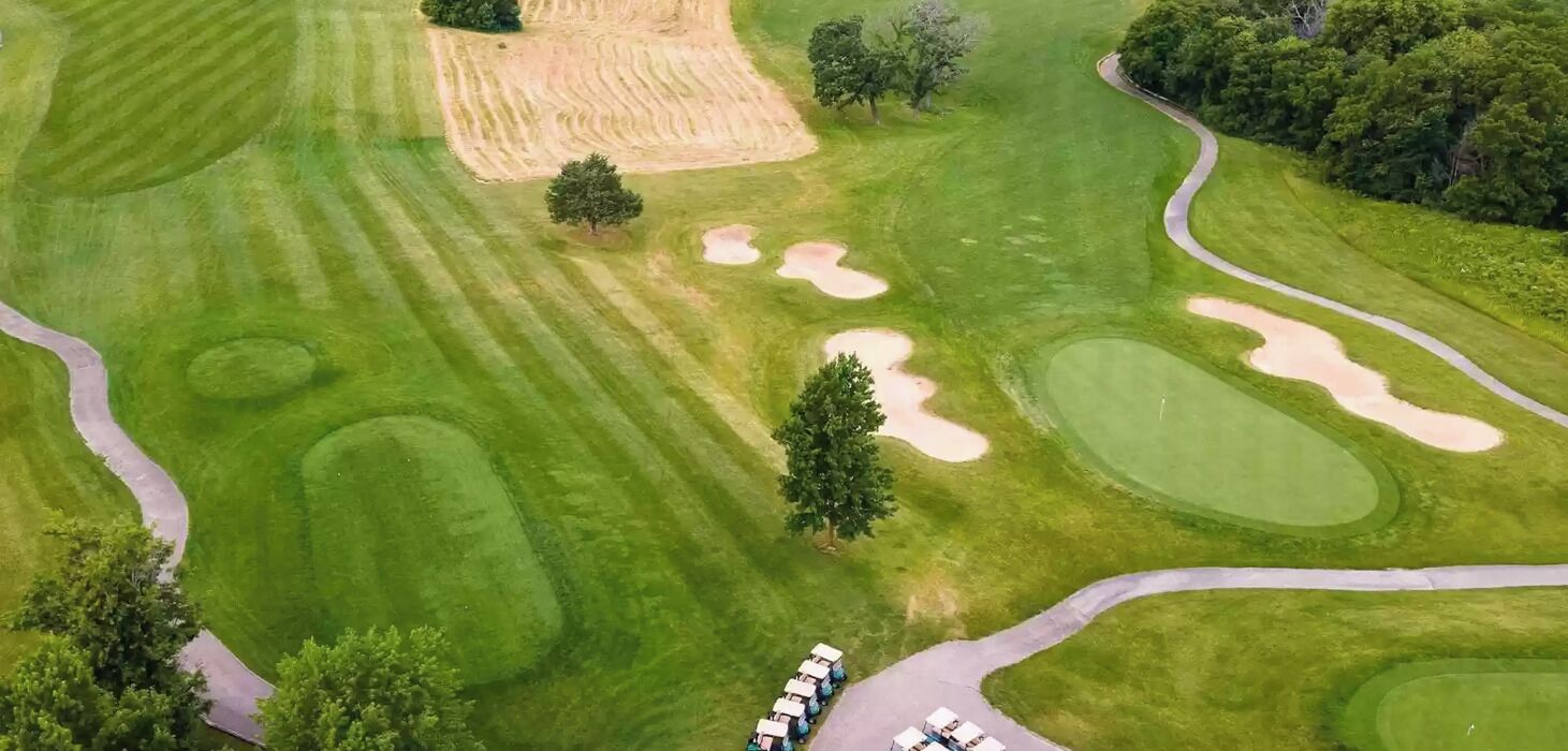 Vista aérea de um campo de golfe com armadilhas de areia, um caminho sinuoso e uma fileira de carrinhos de golfe estacionados.