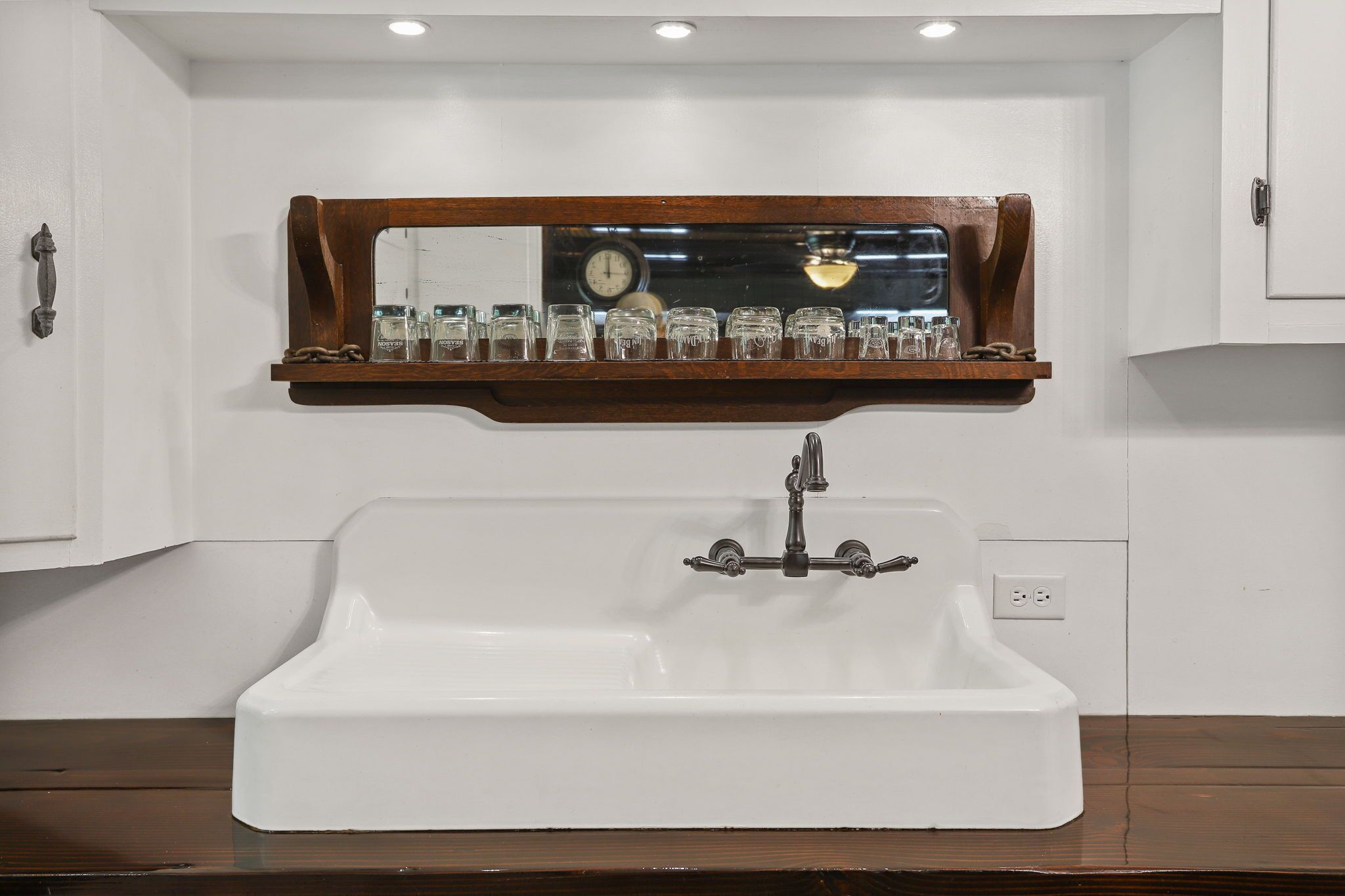 Un lavabo blanc dans une salle de bain, avec une étagère en bois au-dessus, sur laquelle sont posés plusieurs pots en verre et une horloge.
