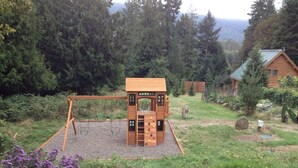 A wooden play structure with a ladder and slides, surrounded by a grassy area with wildflowers and trees. - Cozy St. Helen's Cabin is Pet-friendly, Hot tub, AC, WIFI, Seasonal River Access (Randle)