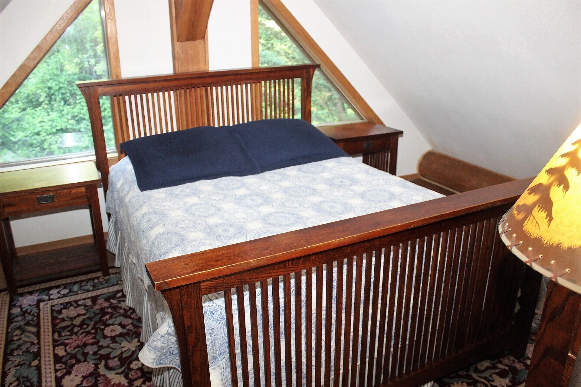 A bedroom with a wooden bed, a nightstand, a lamp, and a view of greenery through a window.