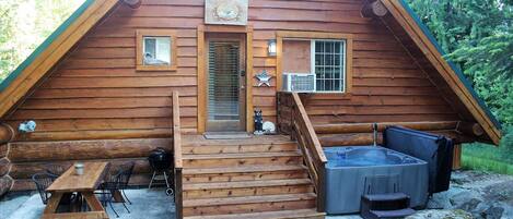A wooden cabin with a porch, a hot tub, and a table with chairs.