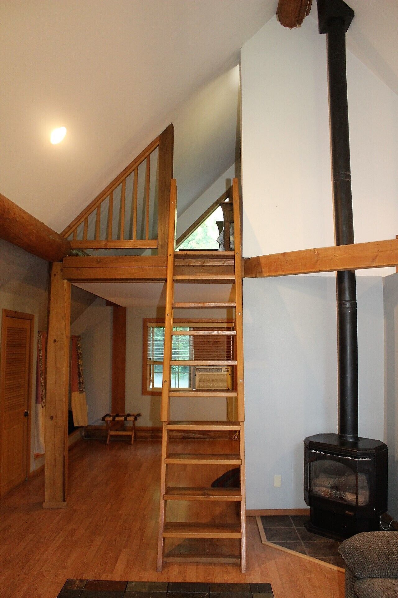 Una escalera de madera que conduce a un dormitorio en lo alto, un estufa de leña negra y una ventana con persianas.