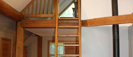 Una escalera de madera que conduce a un dormitorio en lo alto, un estufa de leña negra y una ventana con persianas.