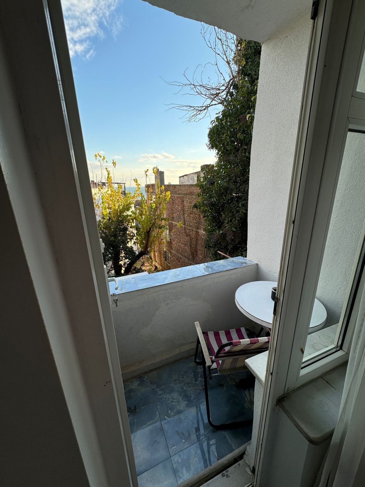 A small balcony with a table and chair, a view of the cityscape, and a door leading to the interior.
