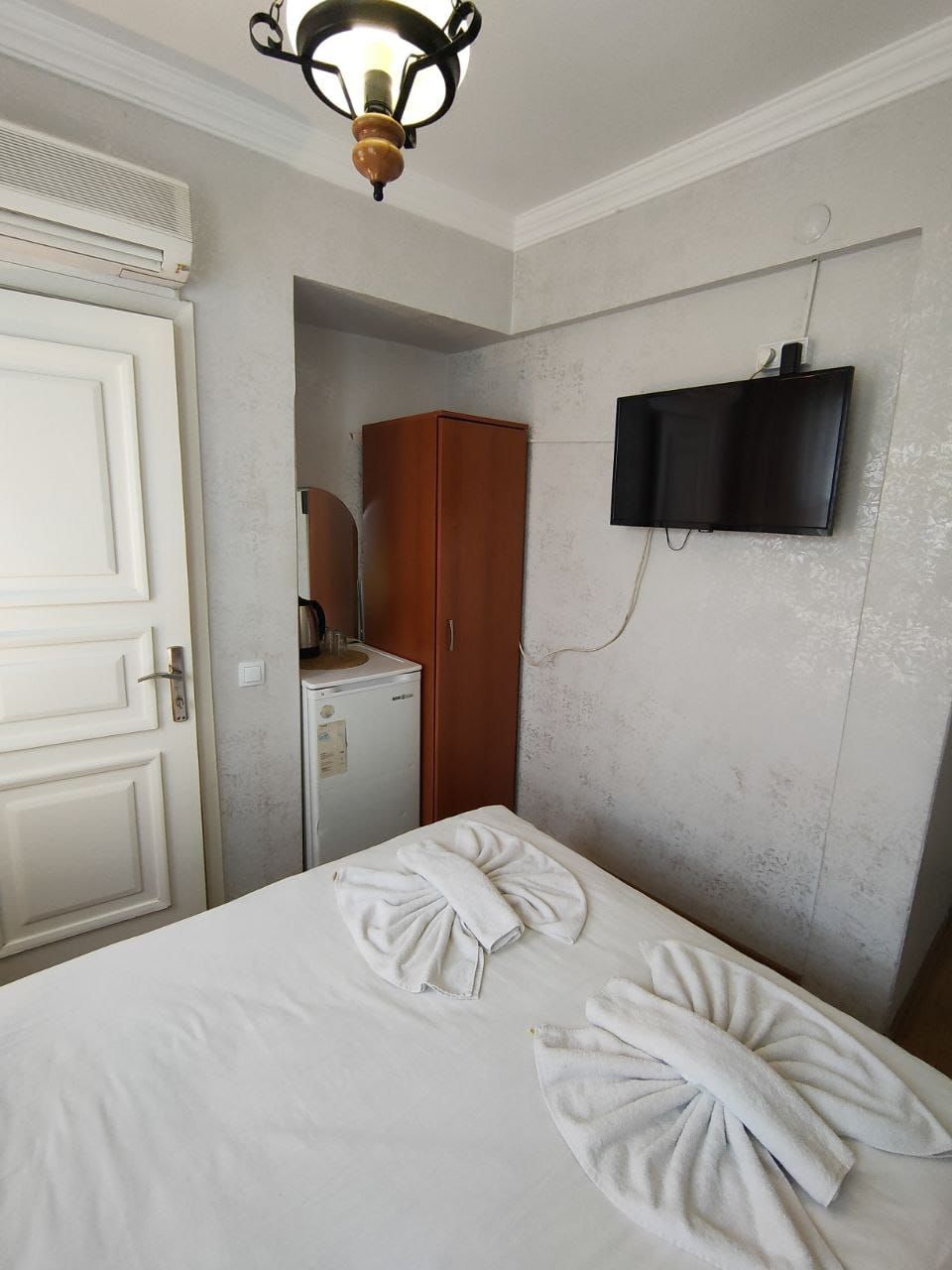 A hotel room with a bed, a wardrobe, a small refrigerator, and a television mounted on the wall.