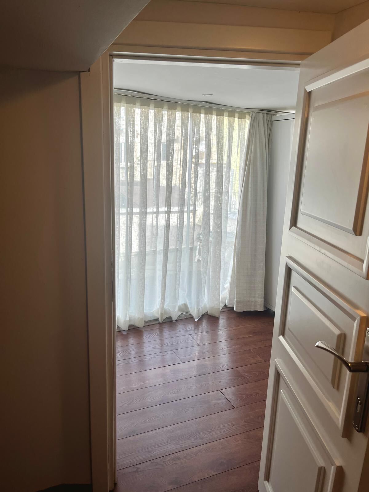 A room with a wooden floor, a white door, and a window with sheer curtains.
