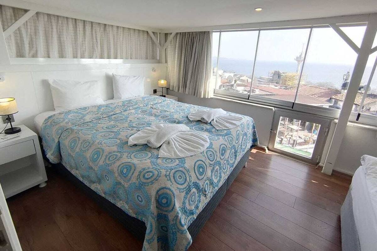 A hotel room with a large bed, a nightstand, a window with a view of the city, and a balcony.