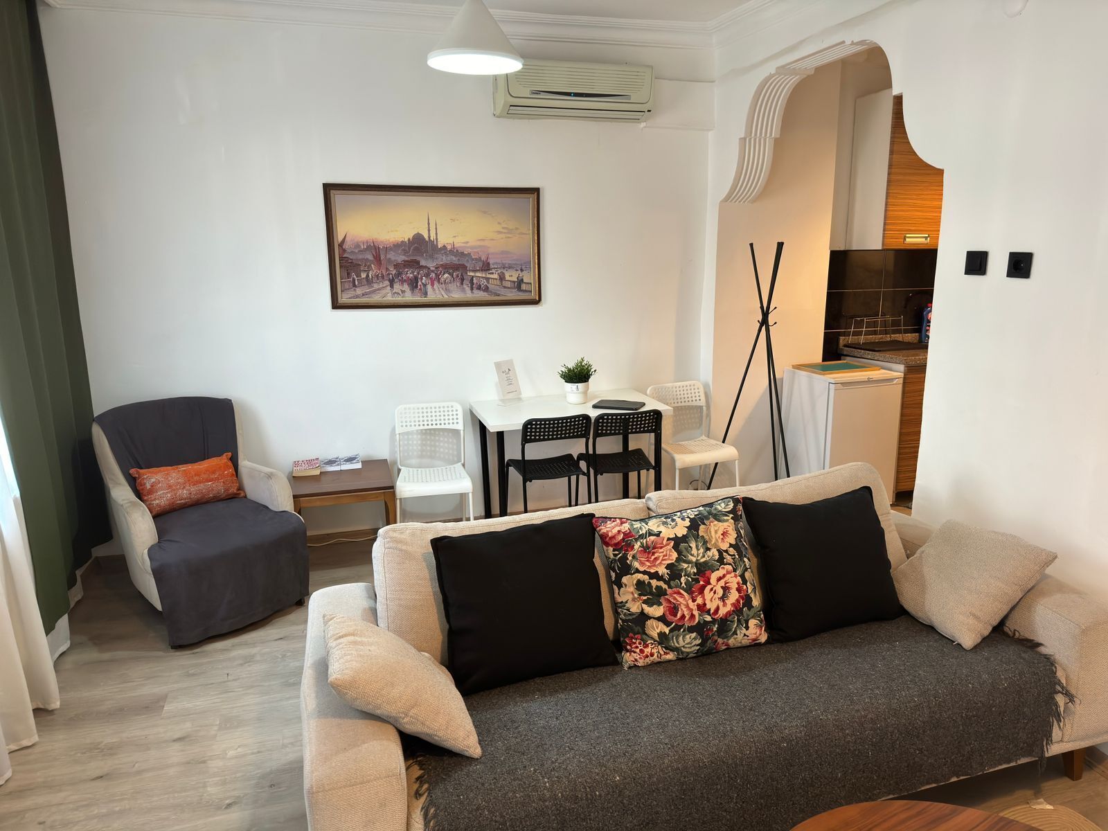 A living room with a sofa, armchairs, a dining table with chairs, a small table with a plant, and a painting of a cityscape.