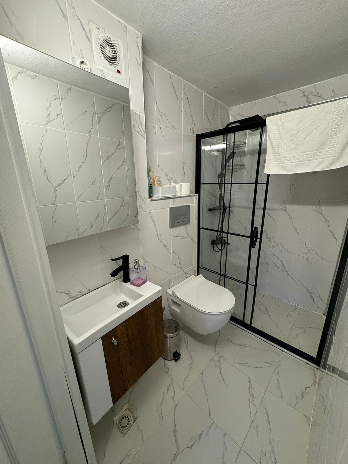 A bathroom with a white marble tile floor, a white toilet, a sink with a wooden cabinet, a glass shower enclosure, and a towel hanging on the shower door.