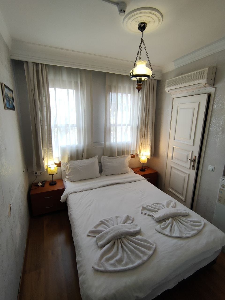 Cama bem arrumada com duas almofadas, mesa de cabeceira com abajur e porta à direita.