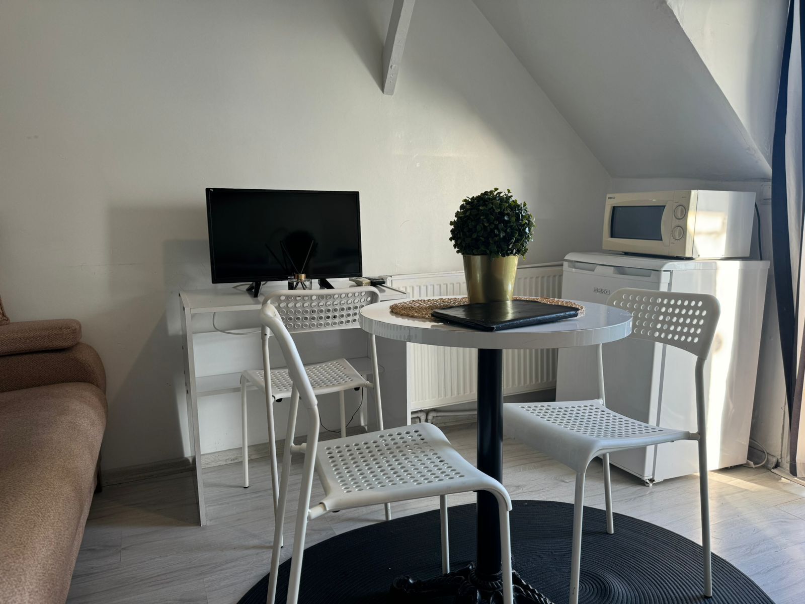A compact living space with a TV, a small table with a plant, two white chairs, and a couch.