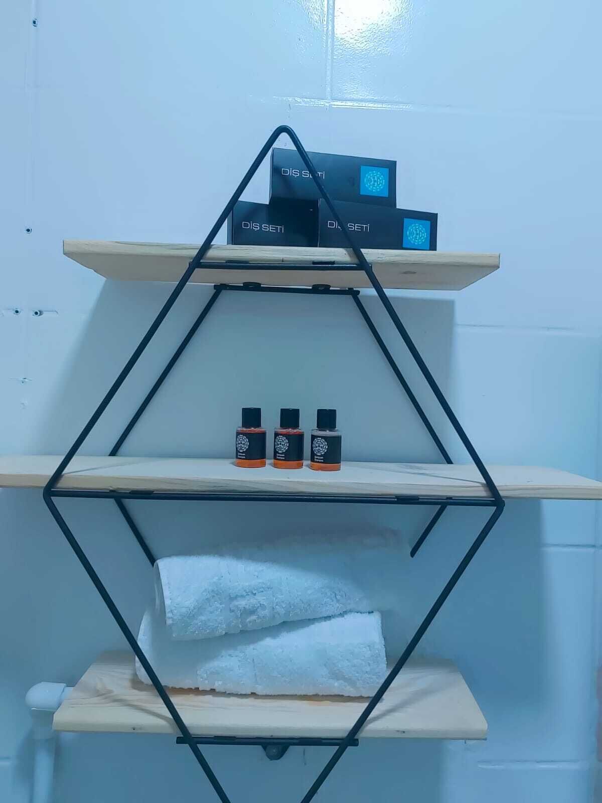 Una estantería metálica de diseño geométrico con tres niveles. El nivel superior contiene dos cajas negras etiquetadas “DIS SETI”. El nivel central tiene tres botellas con tapones naranjas. El nivel inferior contiene una toalla blanca.