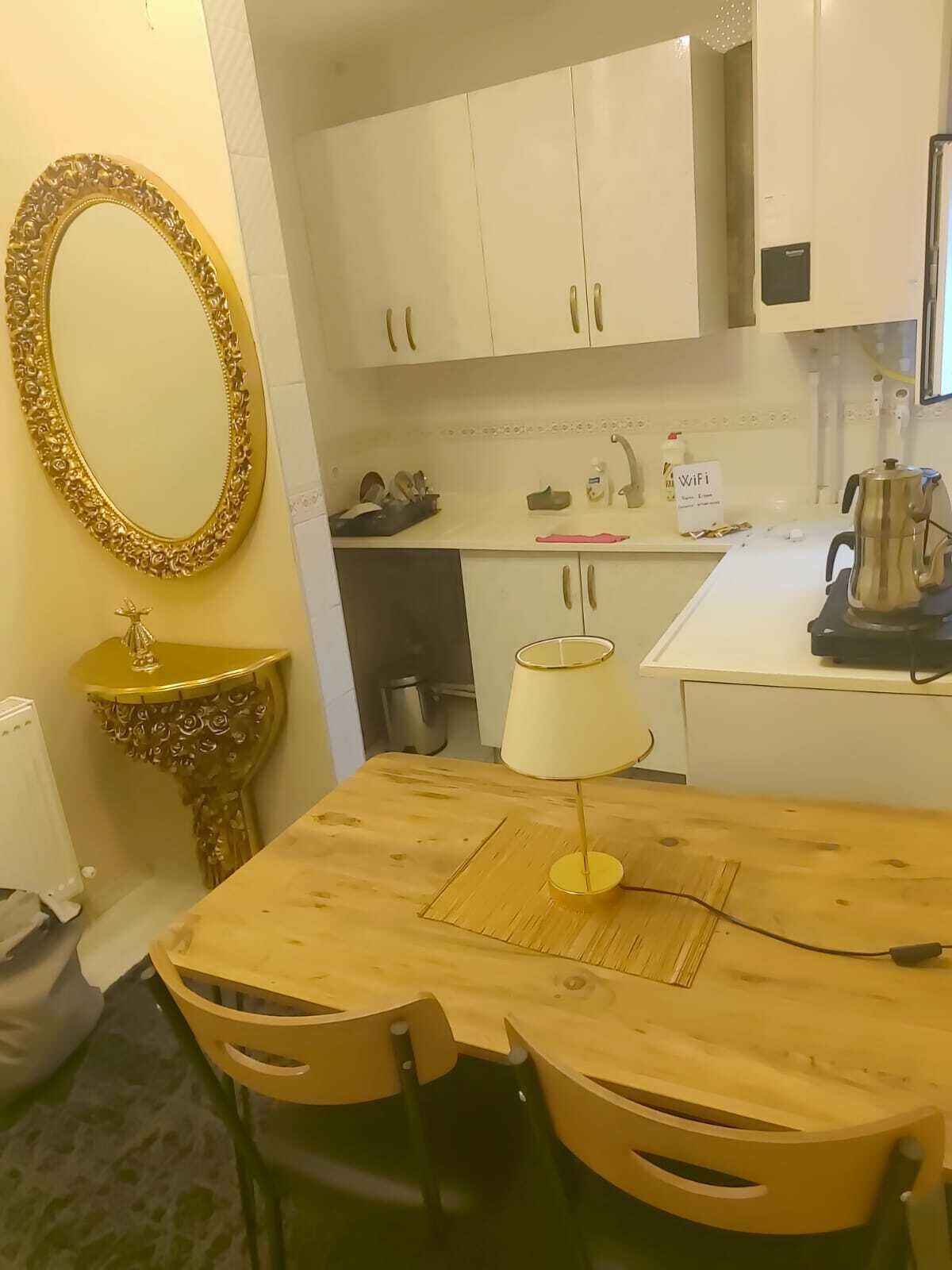 A compact kitchen with white cabinets, a wooden dining table, and a gold-framed mirror.