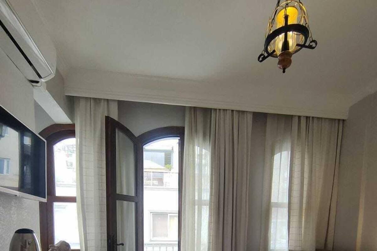 A hotel room with a bed, a desk with a kettle, a chair, and a view through an open door.