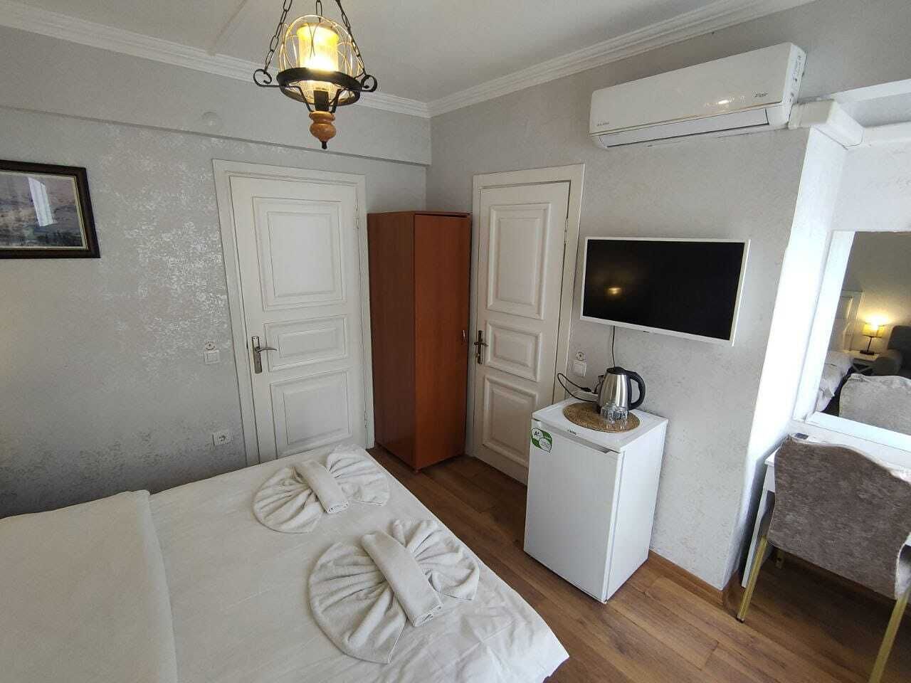 A hotel room with a bed, a desk with a kettle, a television, and an air conditioning unit.