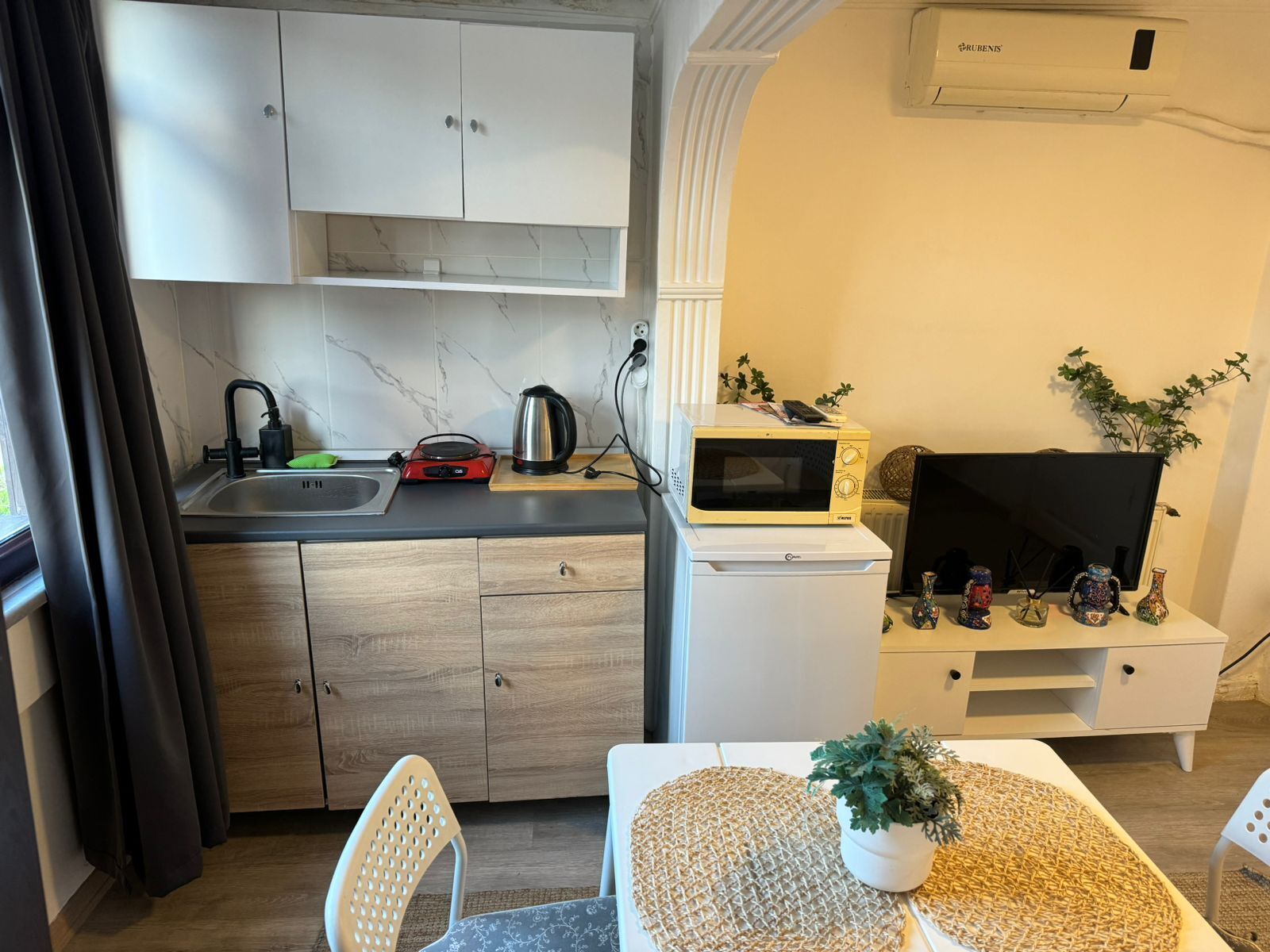 A compact kitchenette with a microwave, kettle, and sink. There is a TV and decorative items on a small table.