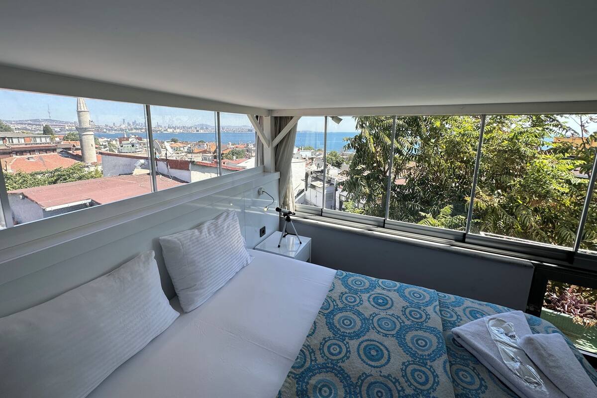 A hotel room with a large window offering a city view, a bed with white linens, and a patterned throw blanket.