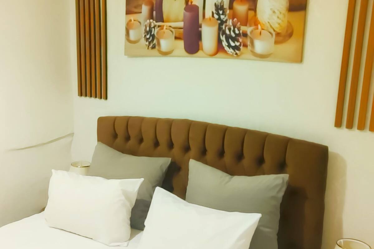 A neatly made bed with white and gray pillows, a brown tufted headboard, and a wall-mounted painting of candles and pinecones.