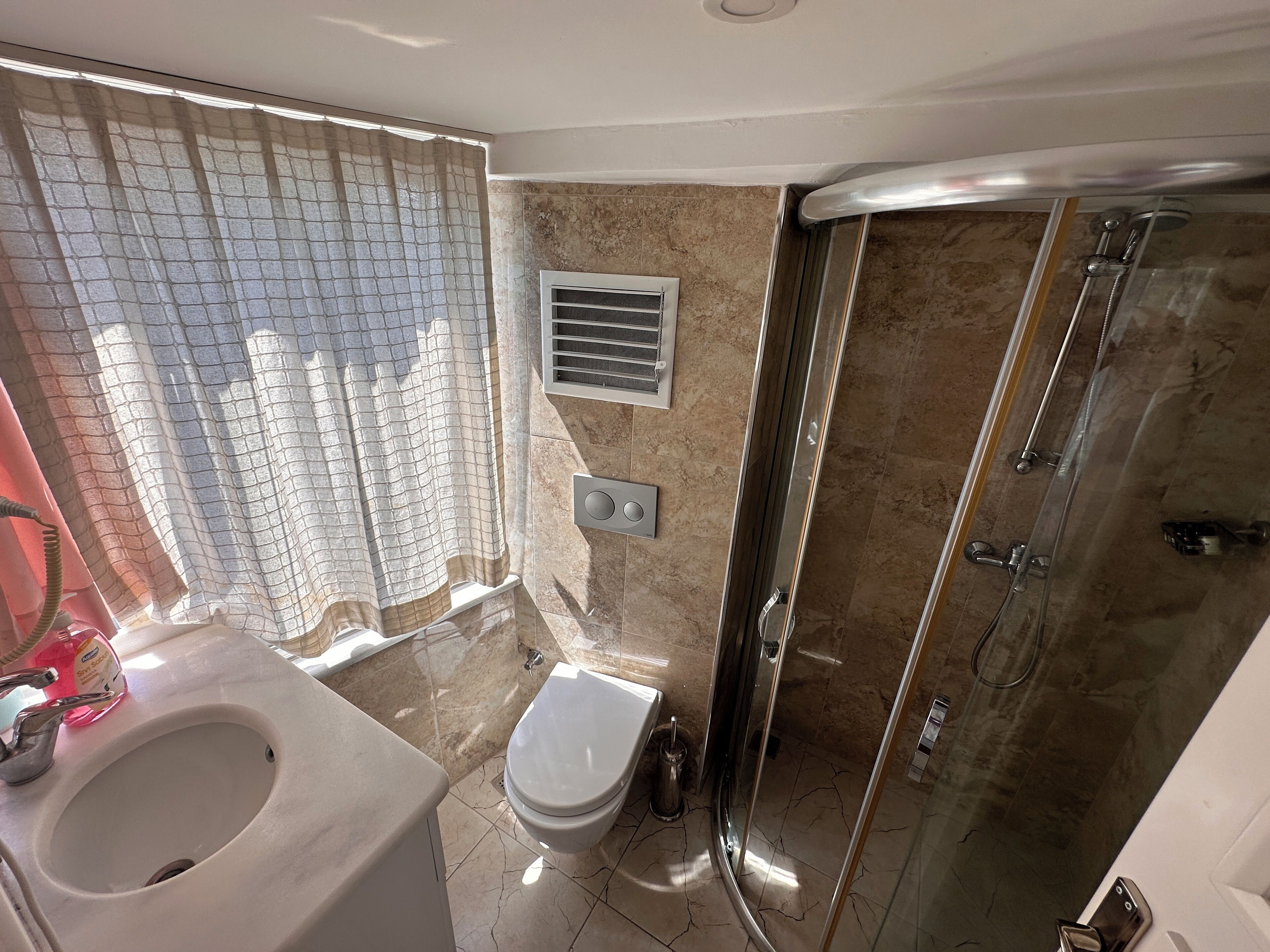 A bathroom with a toilet, sink, shower, and a window with a curtain.