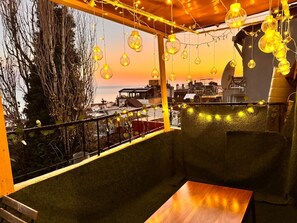 Una terraza en la azotea acogedora con luces esféricas colgantes, una mesa de madera y vistas de la ciudad al atardecer.
