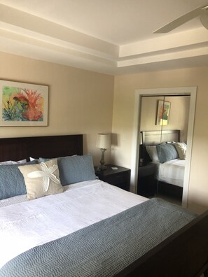 Een hotelkamer met een groot bed, een nachtkastje met een lamp, een schilderij aan de muur en een deur naar een andere kamer.
