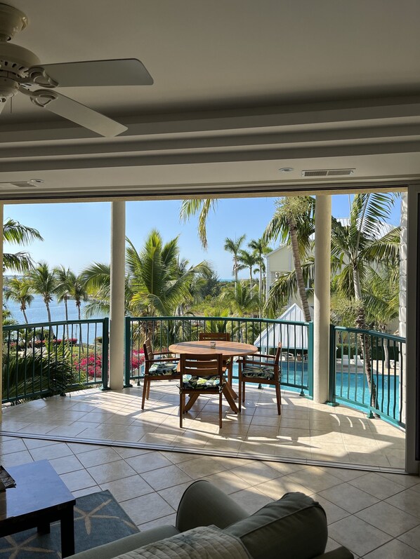 A spacious balcony with a round wooden table and chairs, a view of a swimming pool, and palm trees.