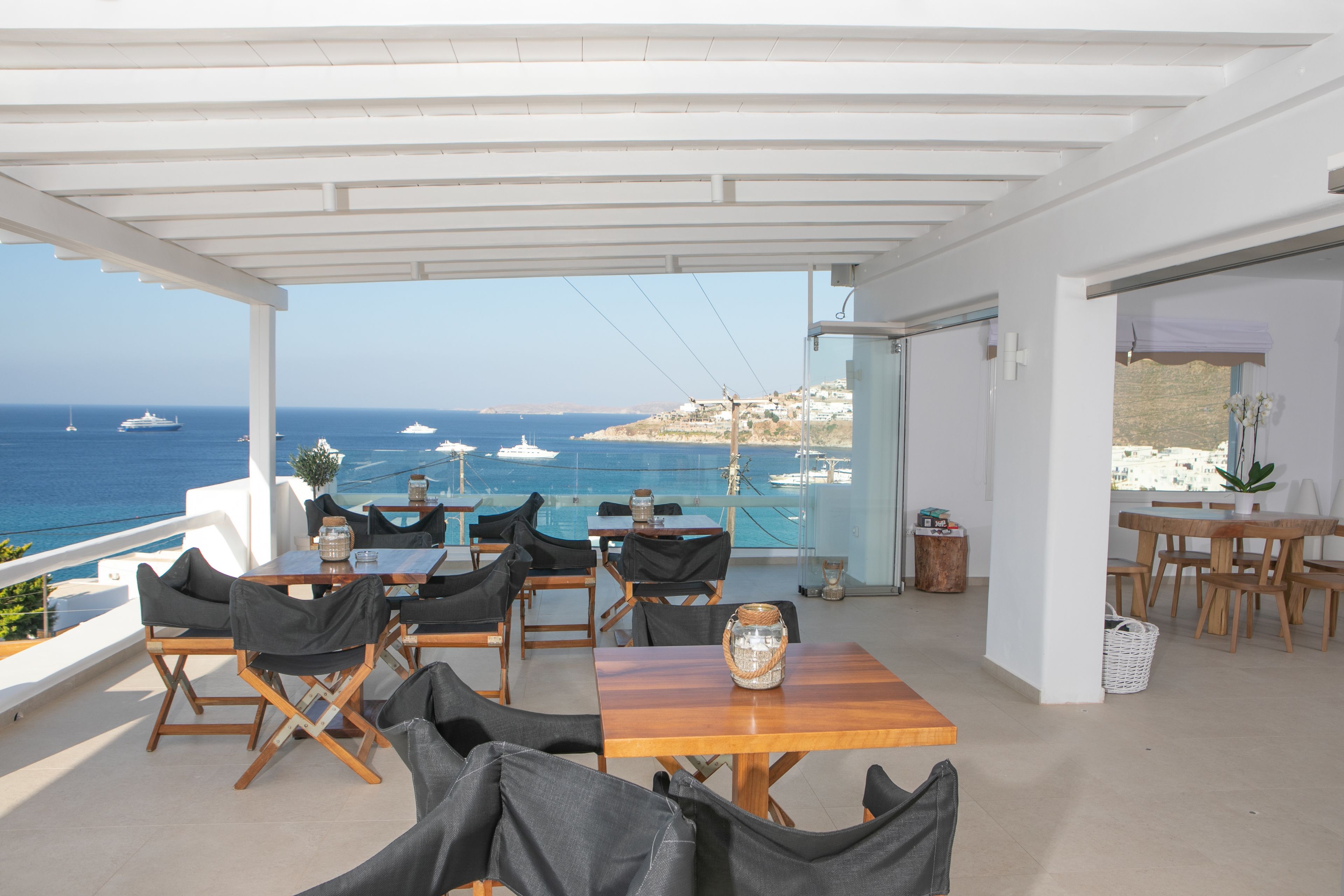 A terrace with tables and chairs overlooking the sea, featuring a clear view of boats and a distant shoreline.