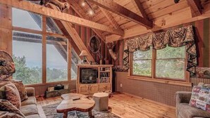 L’intérieur d’un chalet rustique, avec un plafond en bois, de grandes fenêtres, un téléviseur et un coin salon confortable.