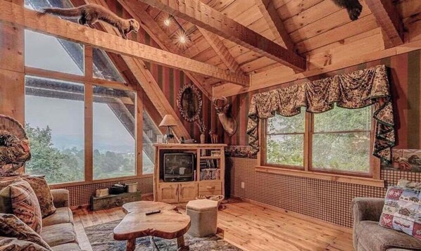 L’intérieur d’un chalet rustique, avec un plafond en bois, de grandes fenêtres, un téléviseur et un coin salon confortable.