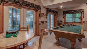 Une pièce chaleureuse équipée d’une table de billard, d’une table de jeu de fléchettes et d’une table à manger. De grandes fenêtres et une porte à panneaux de verre y sont également présentes.