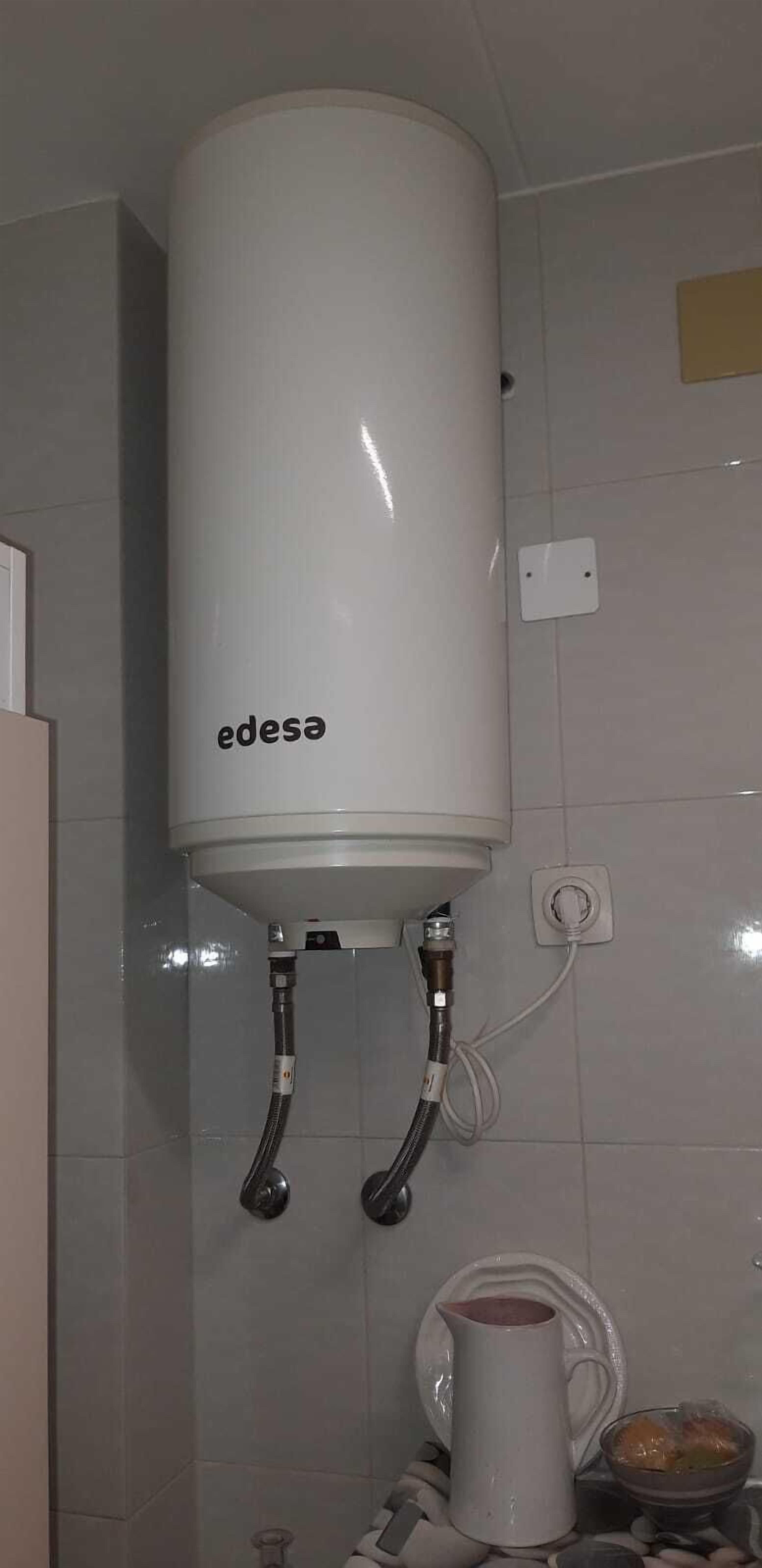 Un chauffe-eau blanc fixé sur un mur recouvert de carreaux, avec deux tuyaux flexibles qui y sont raccordés.