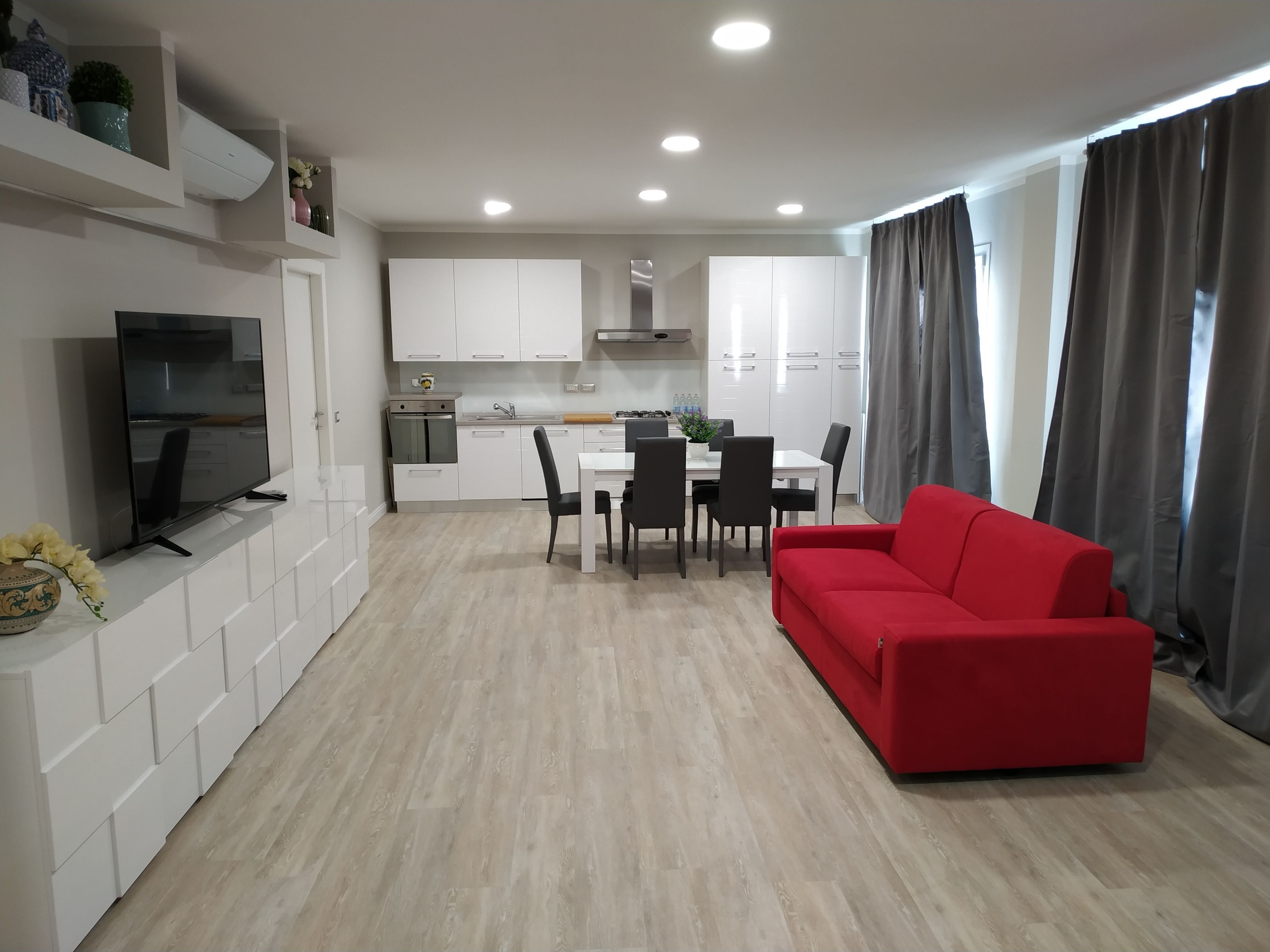 Una sala de estar moderna con un sofá rojo, un comedor con una mesa y sillas blancas, una cocina con armarios blancos y un televisor de pantalla plana sobre un mueble blanco.