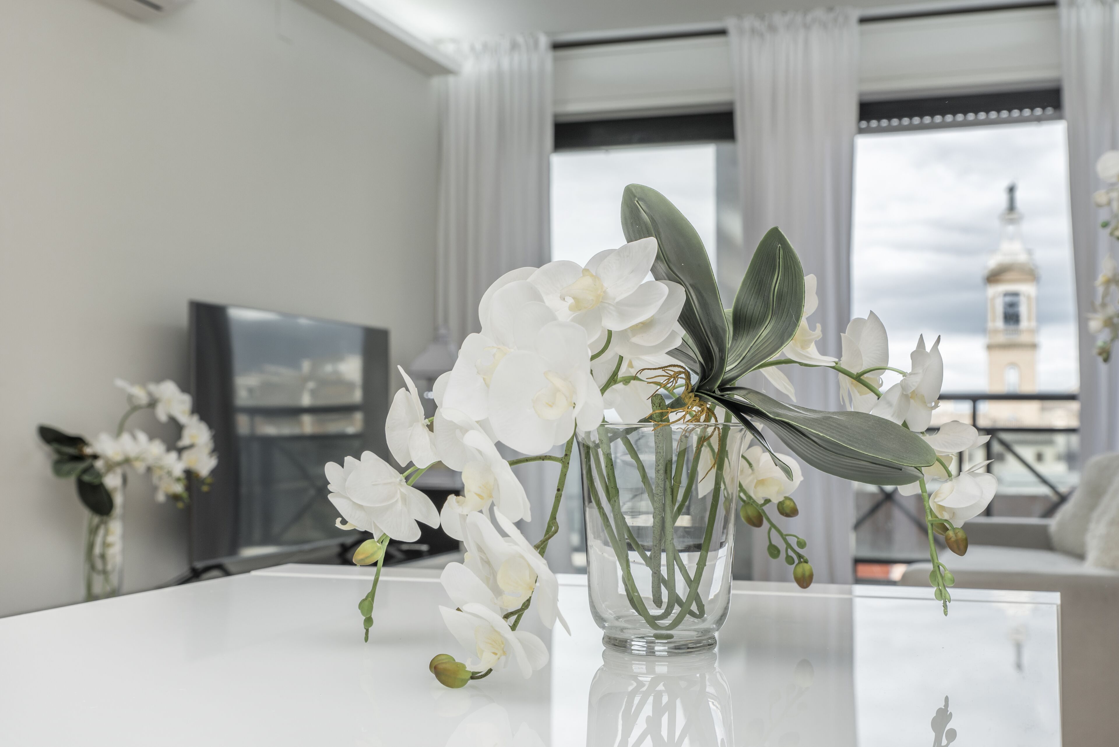 A white orchid in a clear glass vase on a white table in a room with a large window and a view of a tower.