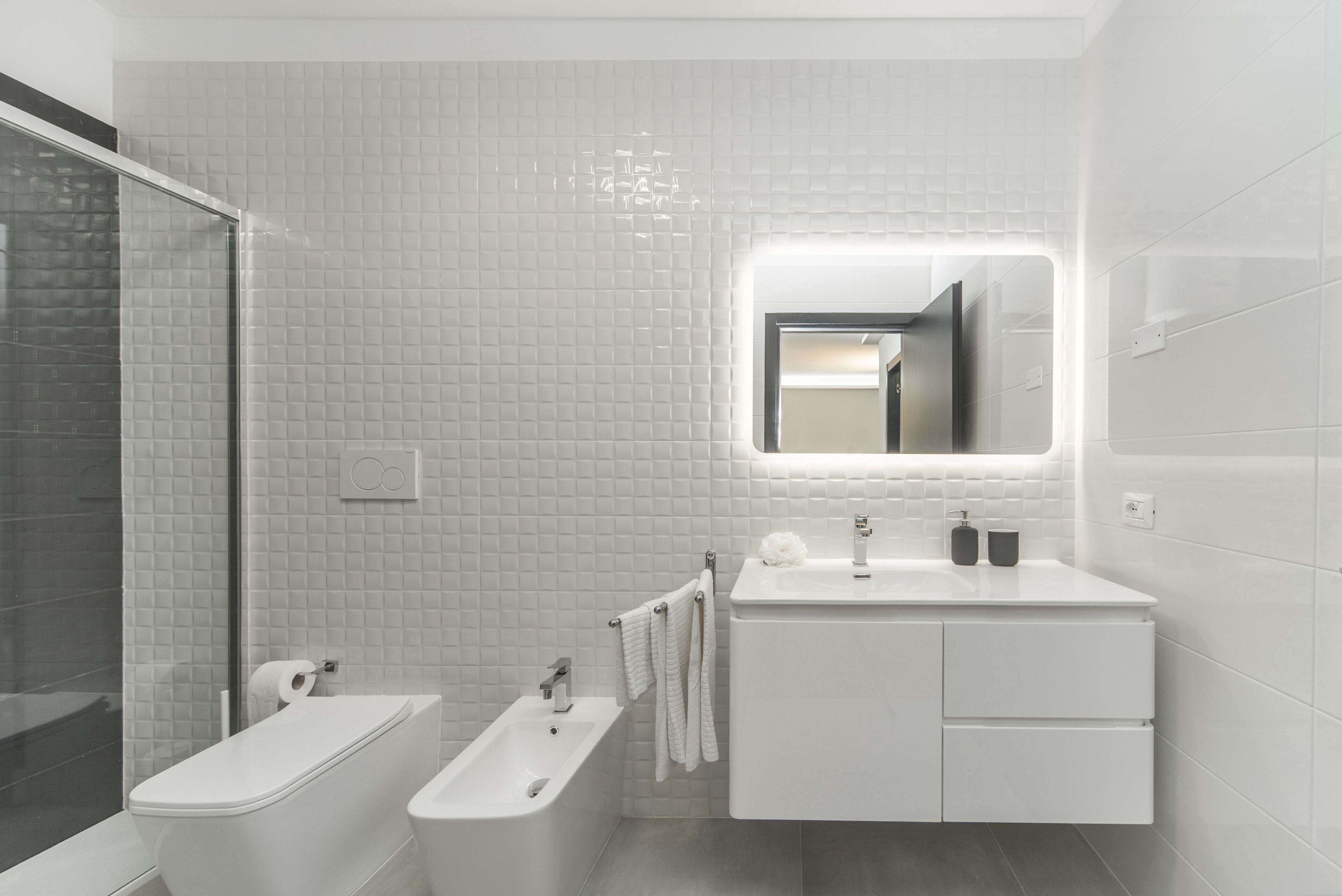 A modern bathroom with a toilet, bidet, and sink, featuring a large mirror and tiled walls.