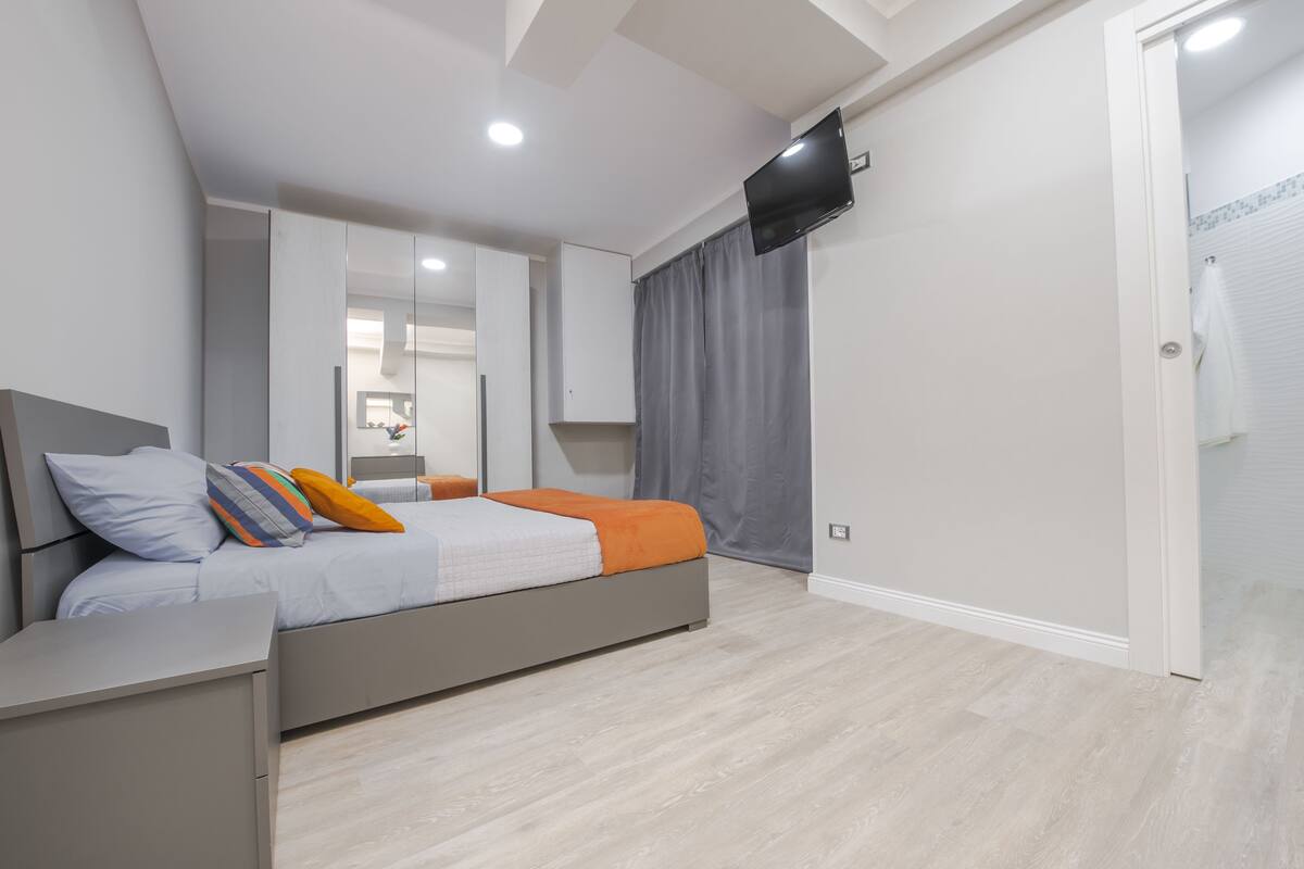 Un dormitorio moderno con cama, mesitas de noche, televisor montado en la pared y puerta que da a un baño.