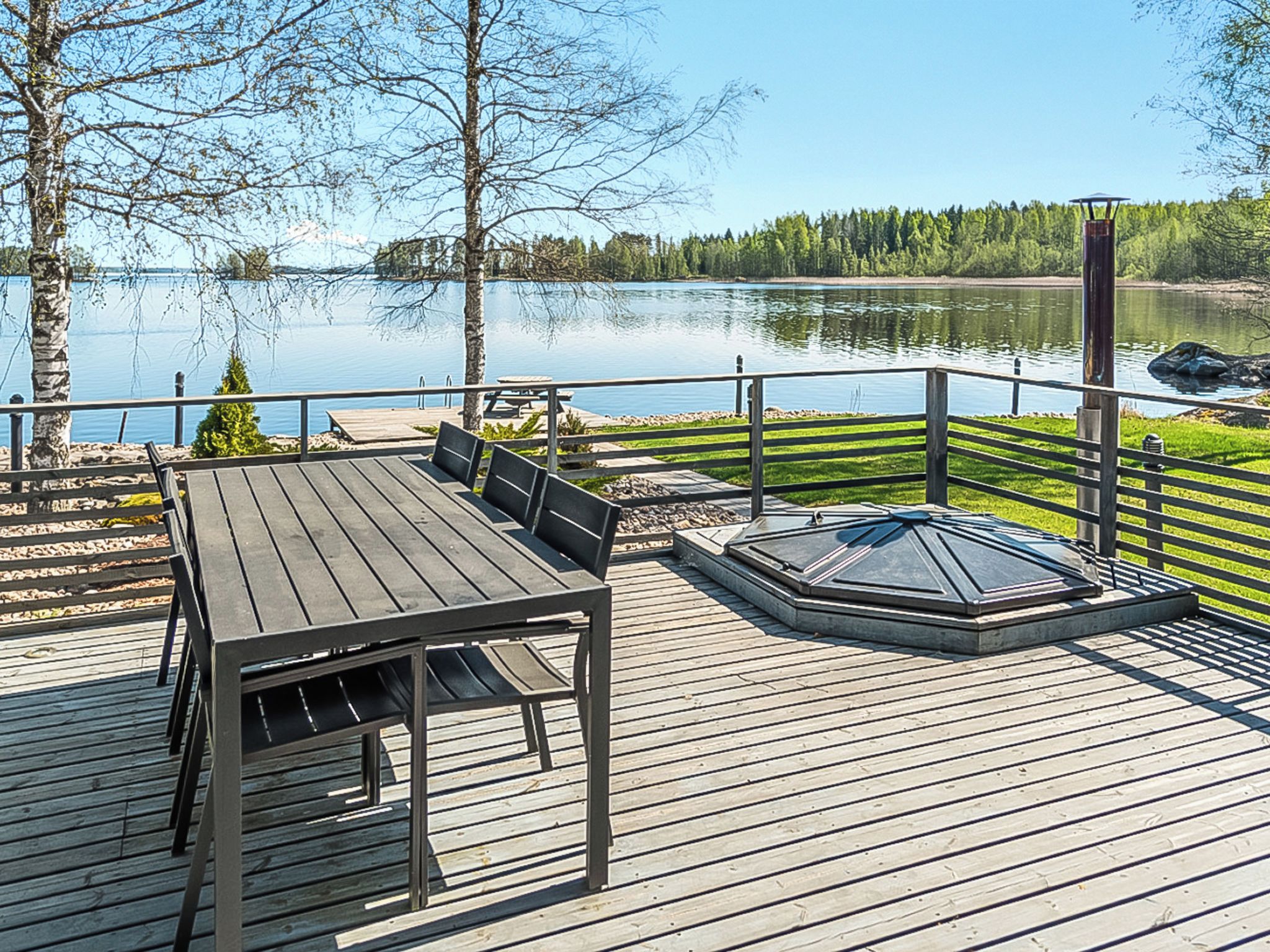 A wooden deck with a table and chairs, overlooking a calm lake with trees in the background.