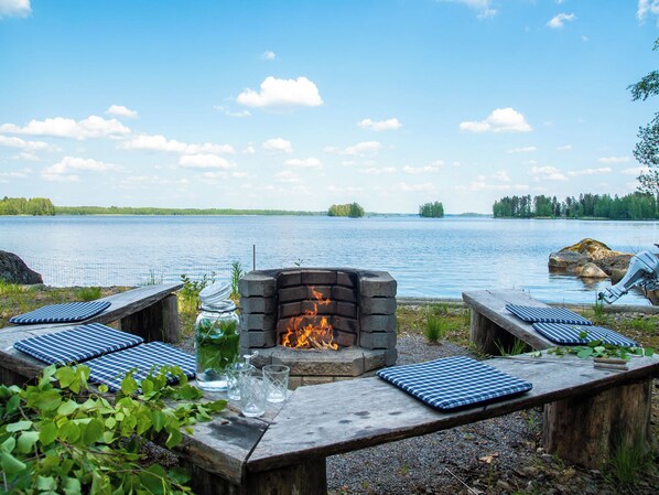 A lakeside outdoor seating area with a stone fire pit, benches with blue cushions, and a fishing rod.