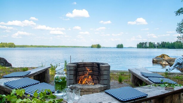 Ein Sitzbereich im Freien am Seeufer mit einem Steinkamin, Bänken mit blauen Kissen und Seeblick.