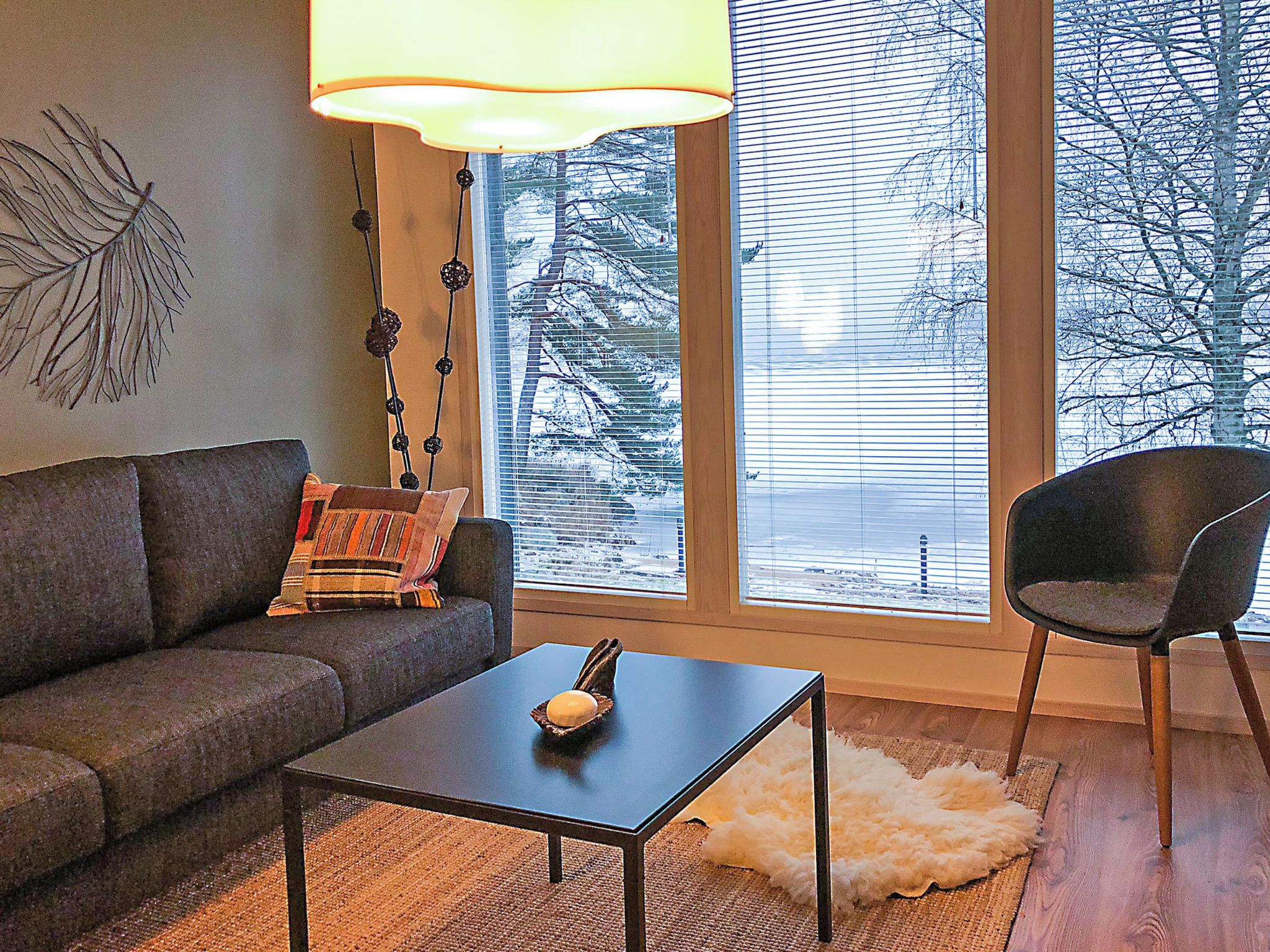 A modern living room with a sofa, a coffee table, and a chair. There is a large window with blinds, a lamp, and a decorative wall piece.