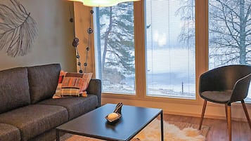 A modern living room with a sofa, a coffee table, and a chair. There is a large window with blinds, a lamp, and a decorative wall piece.