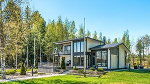 Ein modernes, zweistöckiges Haus mit einer großen Terrasse, umgeben von einem gepflegten Rasen und Bäumen.