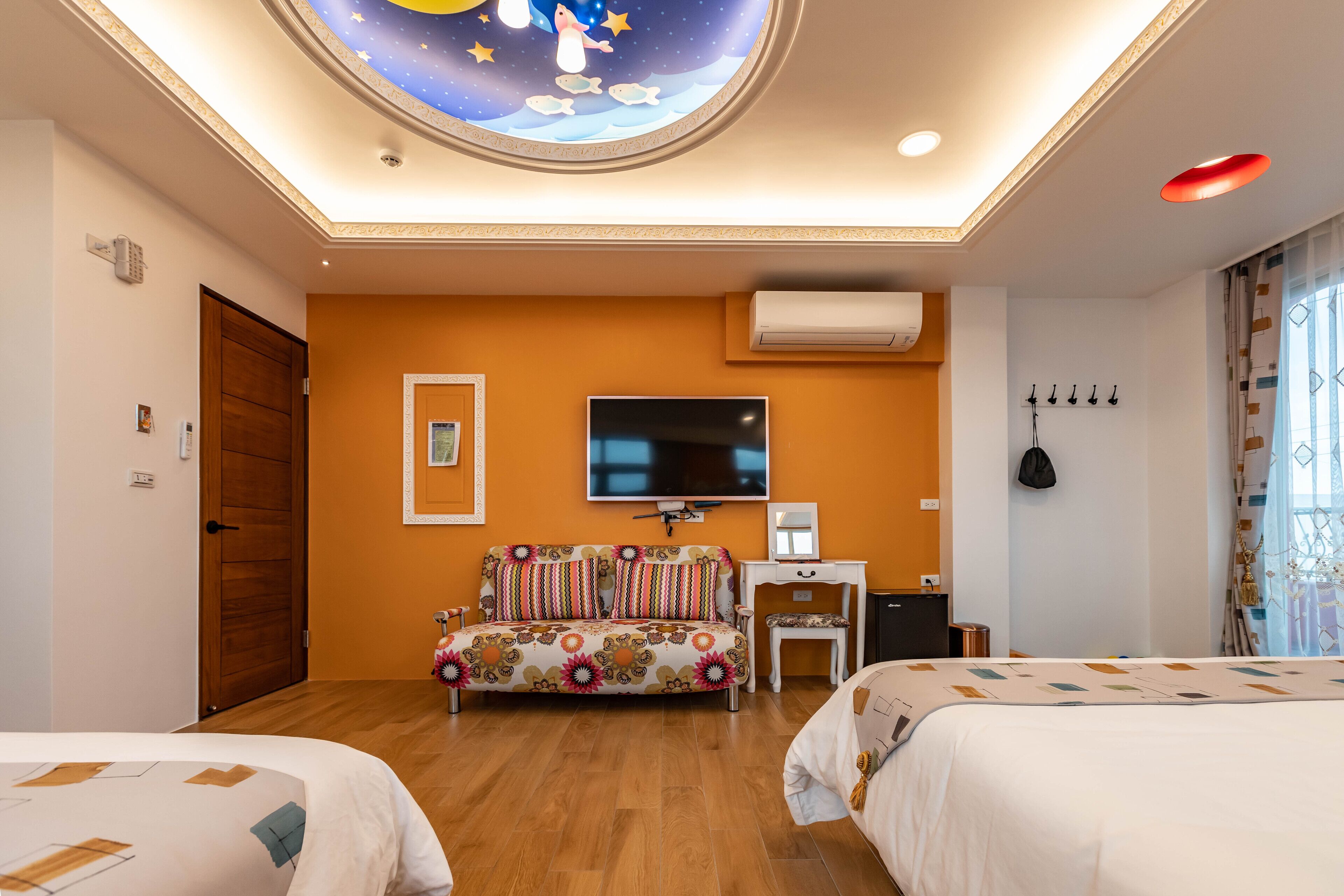 A hotel room with a wooden floor, a sofa with a floral pattern, a flat-screen TV mounted on the wall, and a bed with a geometric-patterned bedspread.
