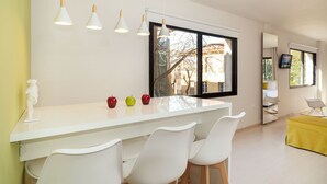 A modern kitchen with a white countertop, three white chairs, and a window with a view of trees and buildings. - Plaka Heart (Athens)