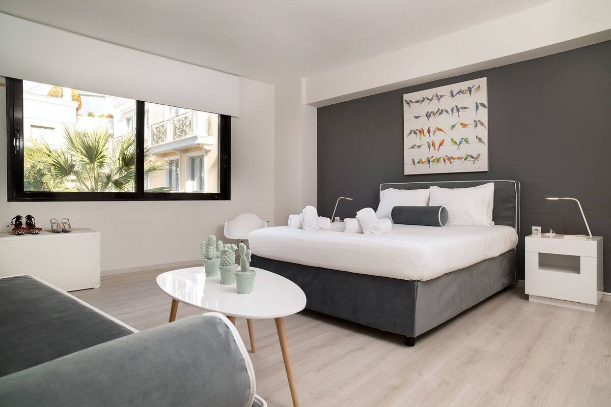 Un dormitorio moderno con una cama grande, una mesita de noche, una mesa redonda y una ventana con vistas a edificios y áreas verdes.