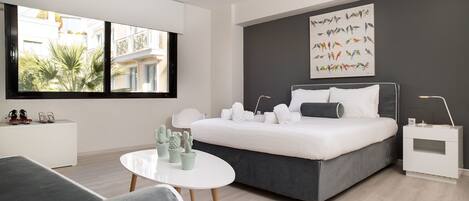 A modern bedroom with a large bed, a bedside table, a round table, and a window with a view of buildings and greenery.