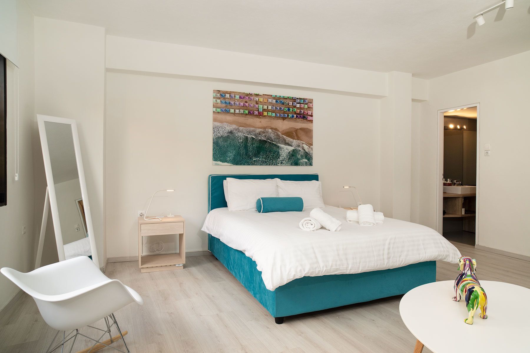 A modern bedroom with a teal bed, white bedding, a large mirror, a small bedside table, and a colorful toy on a round table.