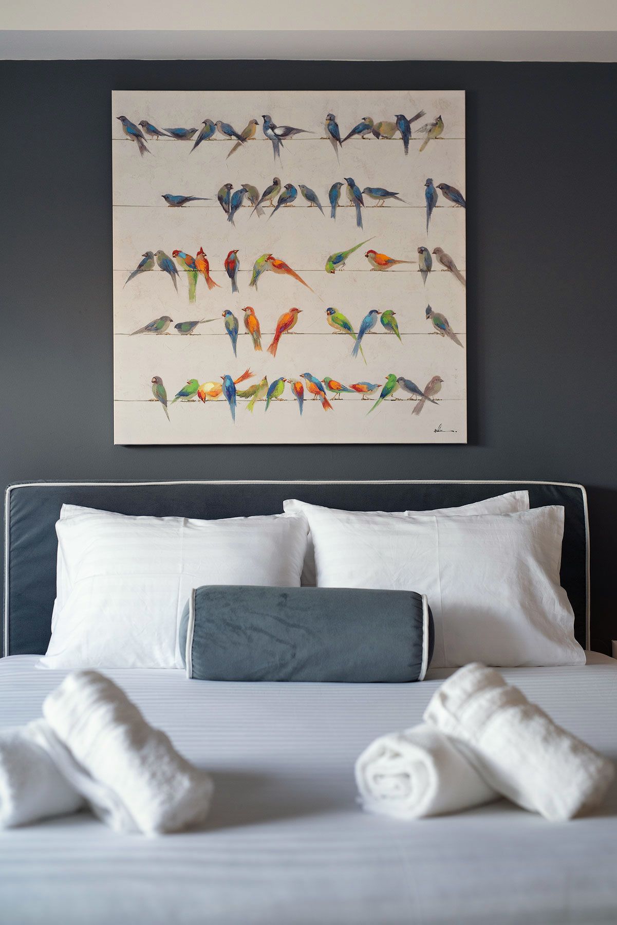 Habitació d'hotel amb un quadre gran de birds de colors a la paret, un llit fet amb roba blanca i un almoiner gris.