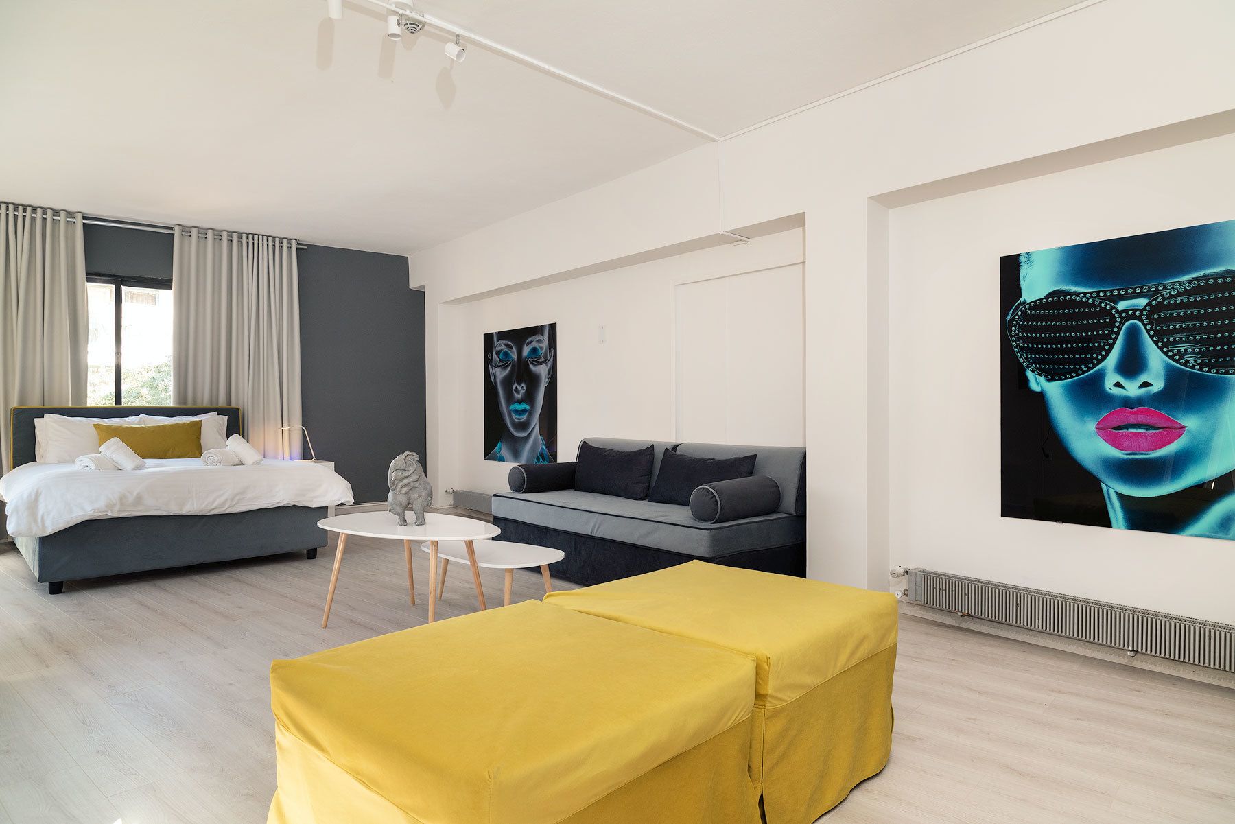 Quarto de hotel moderno com cama, sofá, mesa pequena e um banquinho amarelo. Há duas obras de arte grandes nas paredes.
