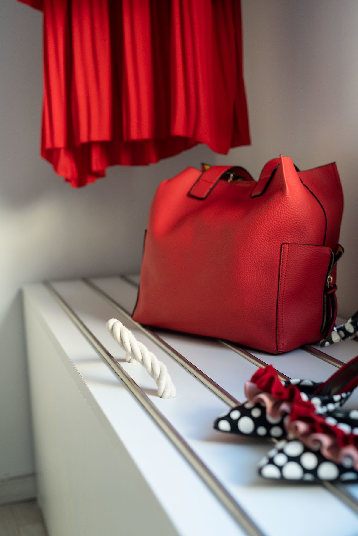 A red handbag on a white bench with a rope and polka dot shoes.