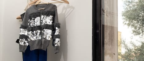 Una habitación con ventana, un perchero de madera con una chaqueta, un bolso azul y un par de zapatillas deportivas sobre un banco blanco.